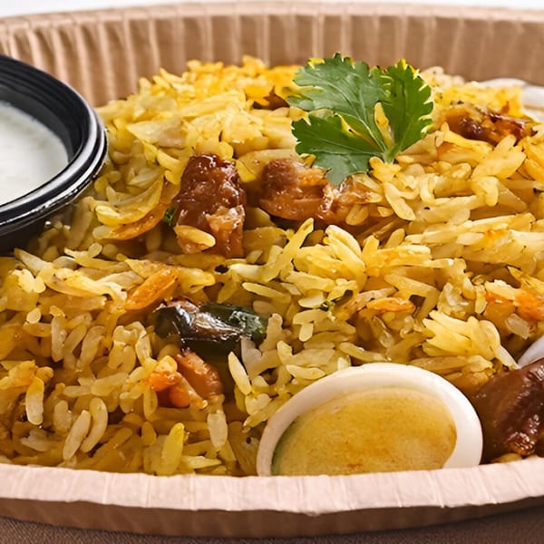 Close-up of Indian biryani with rice, meat, a hard-boiled egg, and a side of yogurt sauce.