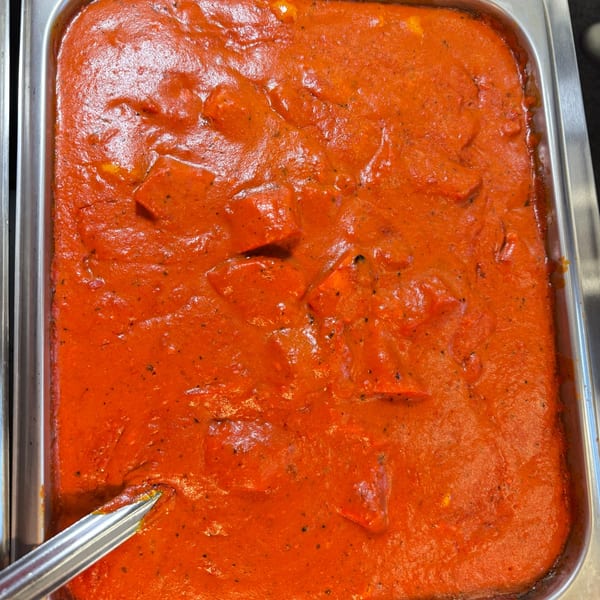 Close-up of a large pan of bright orange Indian curry with paneer, with a serving spoon.