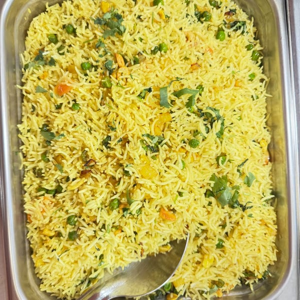 A close-up of yellow Indian rice with green peas, corn, and cilantro in a metal serving dish.